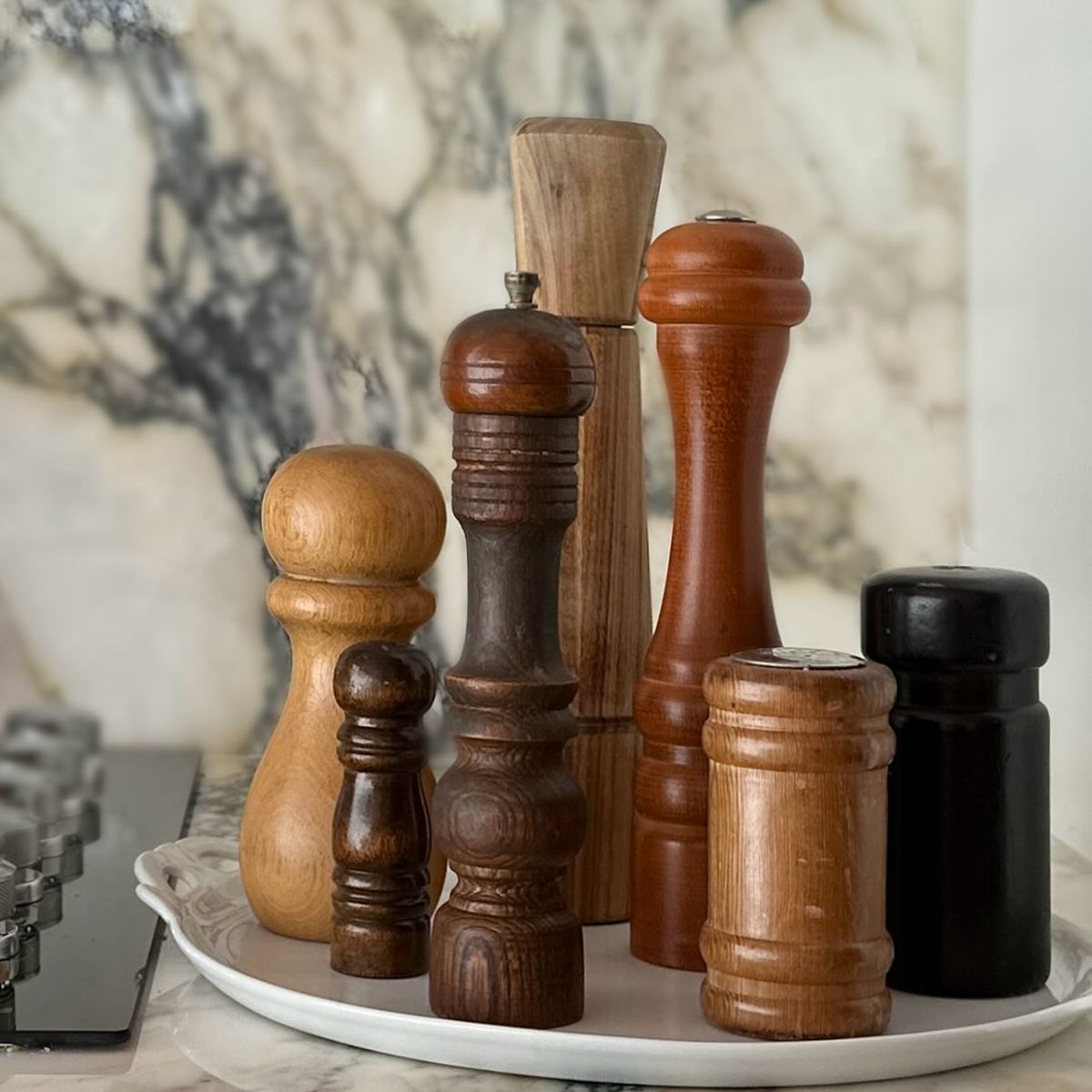 This is an image of a variety of brown wooden salt and pepper shakers and grinders of all sizes on a ceramic white platter with a marble kitchen counter background.
