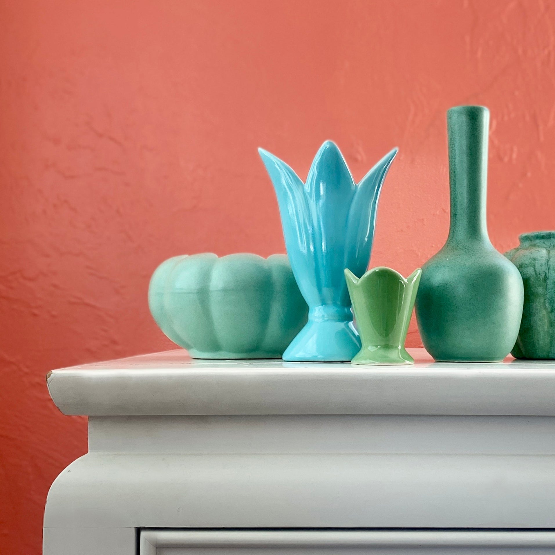 This is an image of five teal ceramic vessels sitting on the corner of a white dresser with a salmon red background. If you click on this image it will take you to our sales platform on Etsy.
