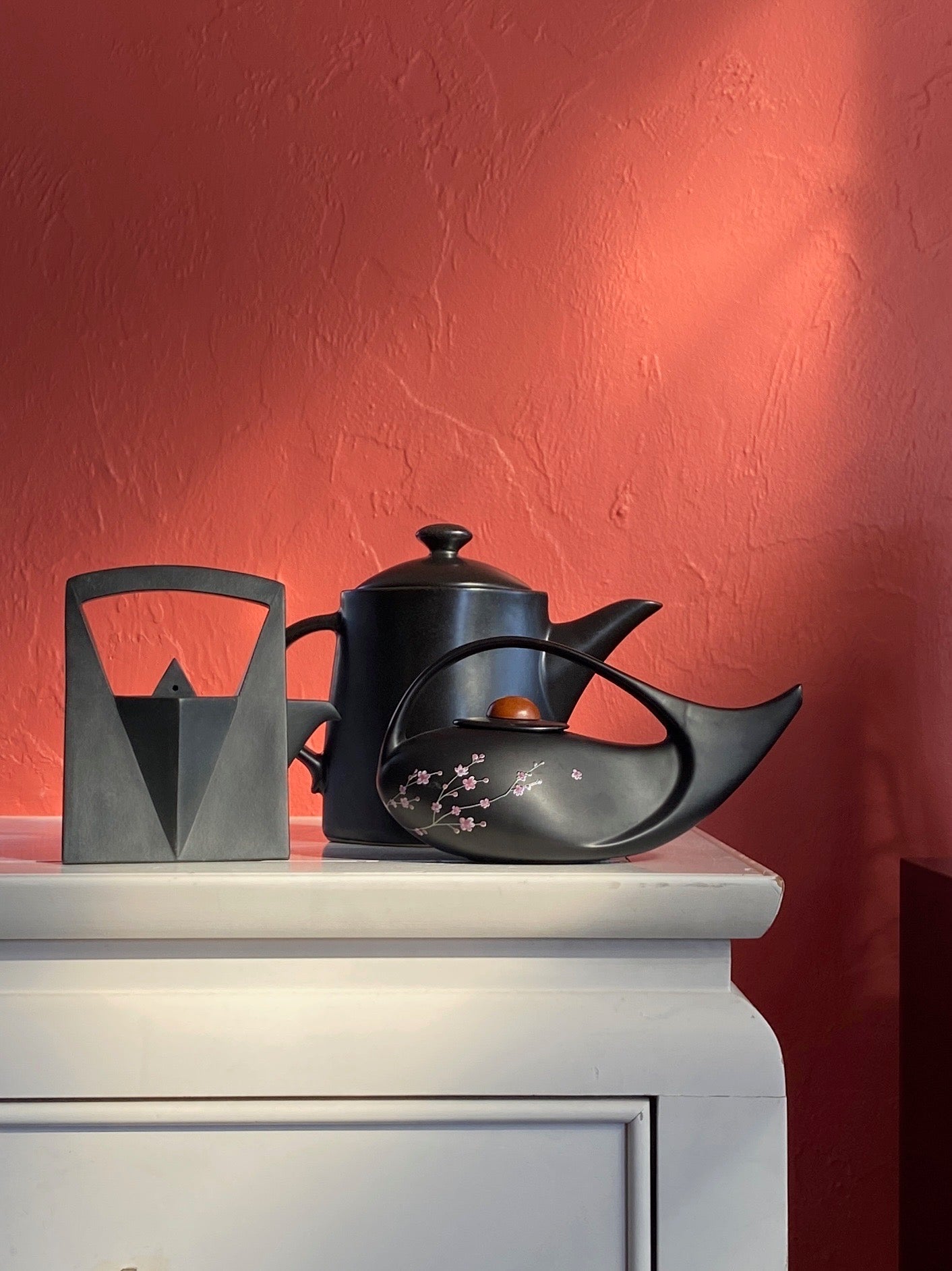 This is an image of three black ceramic teapots sitting on the corner of a white dresser with a salmon red background. If you click on this image it will take you to our sales platform on Chairish.
